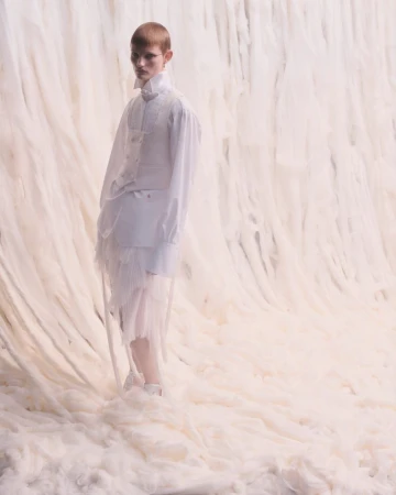 Fashion shot of a person in a white outfit on a soft, fluffy white surface against a draped backdrop, fashion-forward editorial style.