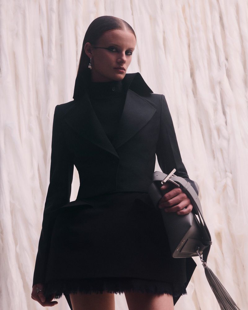 Model wearing a structured black tailored jacket and holding a grey tassel handbag in front of a white fringed textured backdrop.