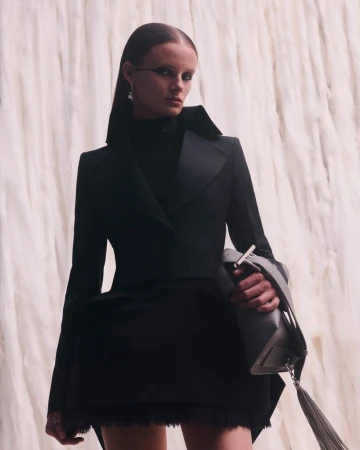Model wearing a structured black tailored jacket and holding a grey tassel handbag in front of a white fringed textured backdrop.