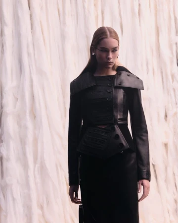 Model wearing a structured black leather jacket and skirt against a draped white textured textile backdrop.