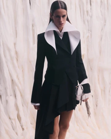 Model wearing a black jacket with a large white collar and holding a light-colored handbag against a textured white background.