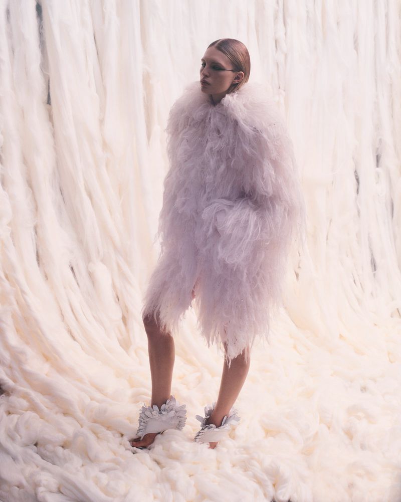 Model wearing an oversized pale textured feather coat and ruffled heels standing against a cascading white wool backdrop.