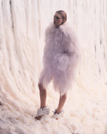 Model wearing an oversized pale textured feather coat and ruffled heels standing against a cascading white wool backdrop.