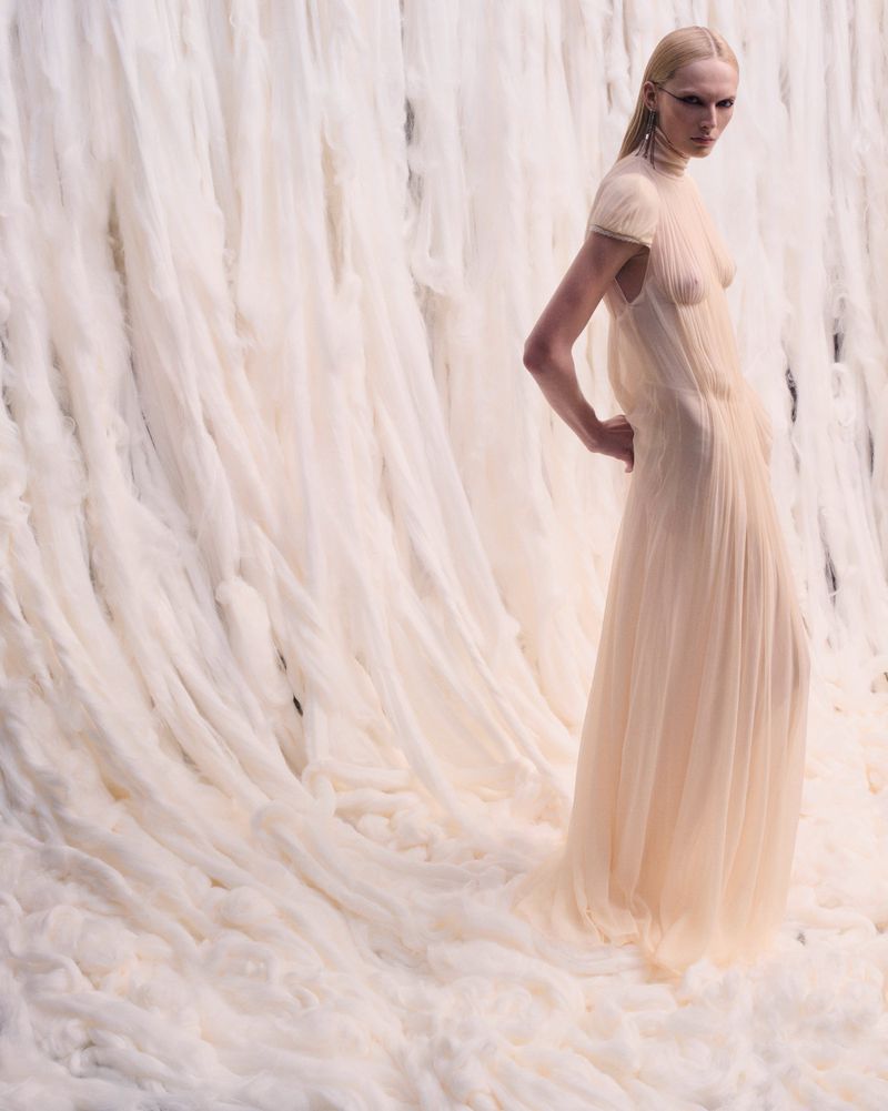 Fashion shot of a slim, fair-skinned adult female model with minimalist makeup, posing in a draped ivory gown, set against an off-white draped background.