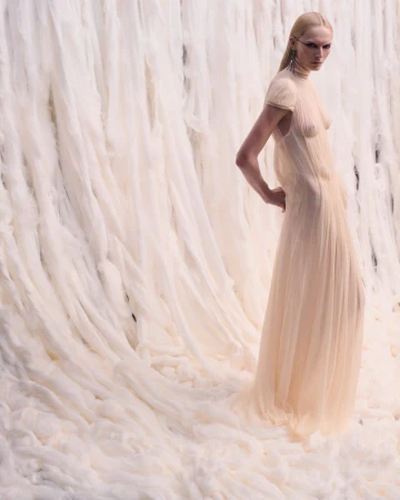 Fashion shot of a slim, fair-skinned adult female model with minimalist makeup, posing in a draped ivory gown, set against an off-white draped background.