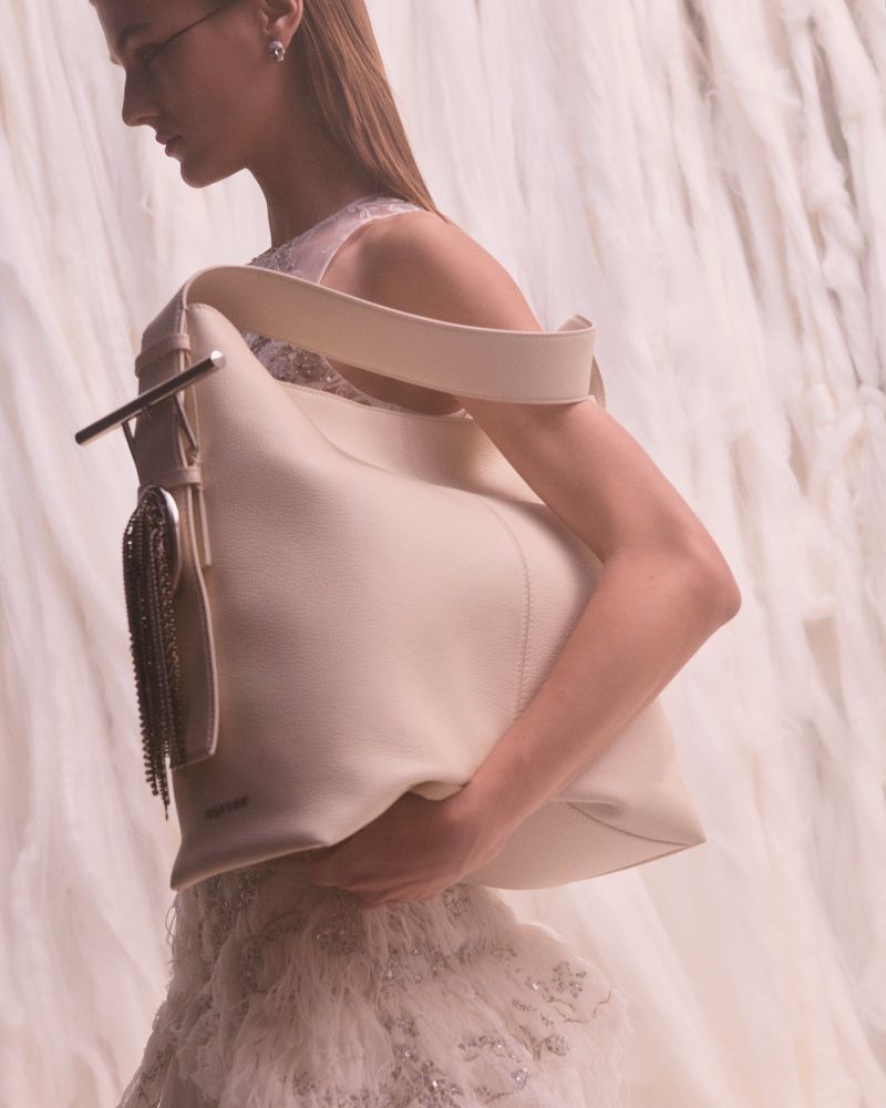 Woman holds a large, cream-colored leather handbag with a chain tassel against a backdrop of soft, draped white fabric.