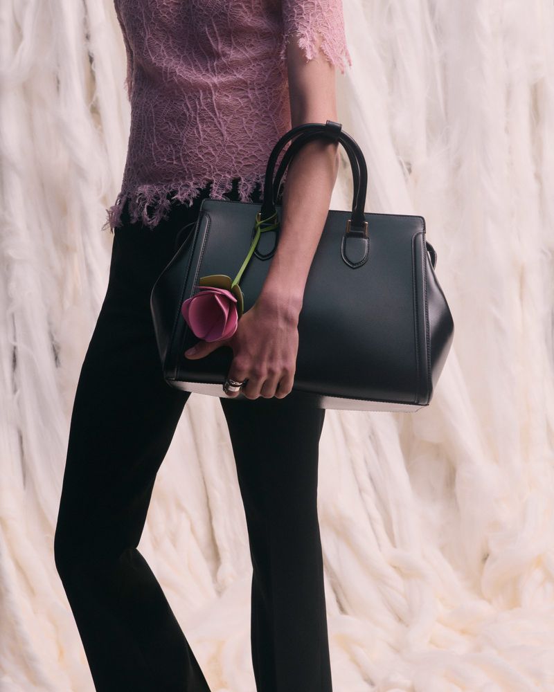 Black leather handbag with a rose charm held by a person wearing a pink lace top and black pants in a neutral backdrop.