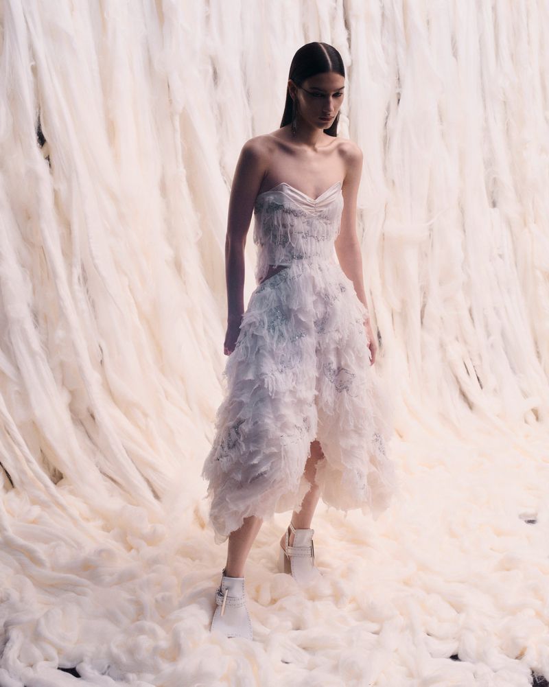 A female fashion model in a white layered dress and white boots poses against an off-white draped fabric backdrop in a studio setting.