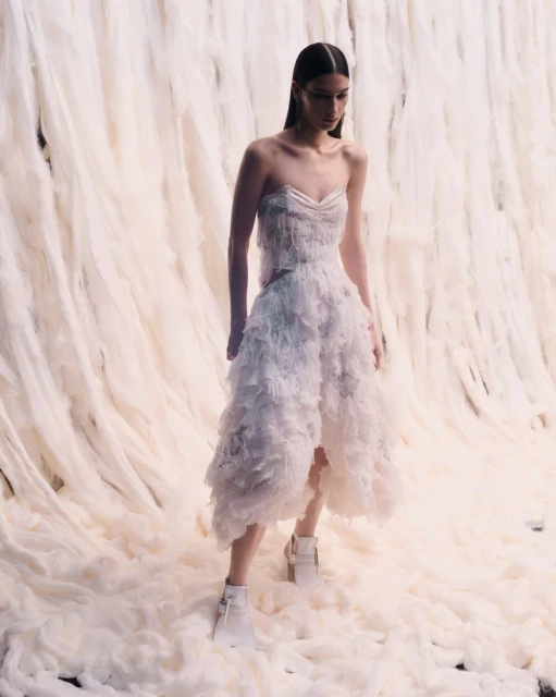 A female fashion model in a white layered dress and white boots poses against an off-white draped fabric backdrop in a studio setting.