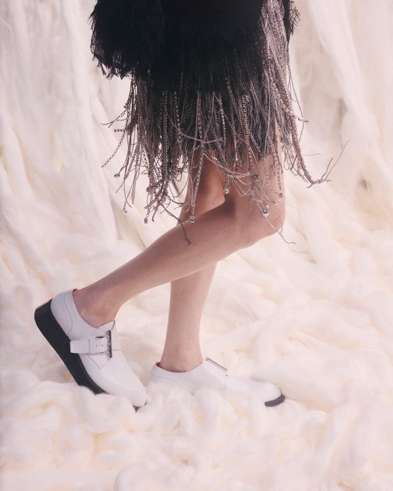 Model's legs and white buckle shoes stand on fluffy fabric. The model wears a black skirt with a fringe of hanging silver chains.