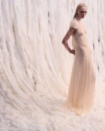 Model with slicked blonde hair in a sheer cream gown standing against a textured raw fiber wall for Alexander McQueen.