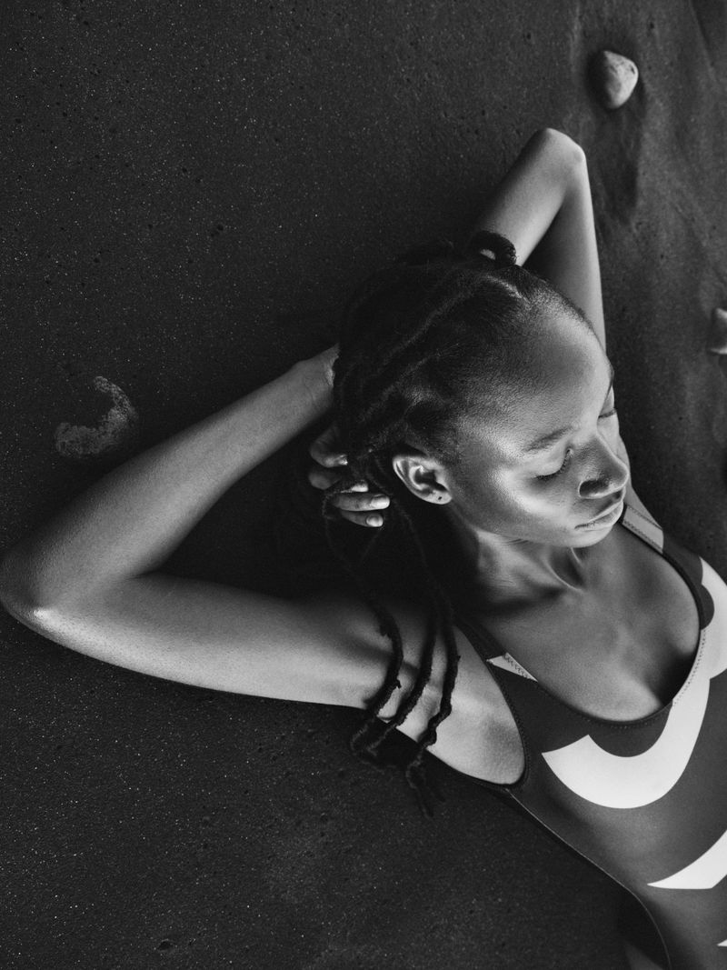Monochrome portrait of a black woman with braids relaxing on a black sand beach in a patterned bathing suit, eyes closed in daylight.