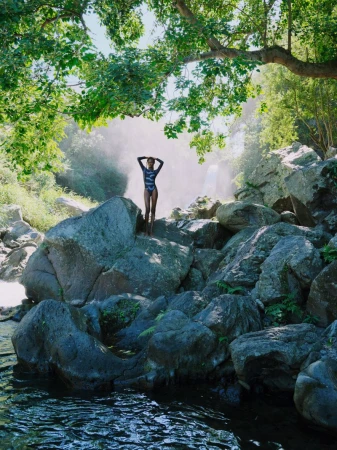 Woman in blue swimsuit standing on rocks near waterfall with forest backdrop on a bright day, hands on head. Outdoor nature setting.