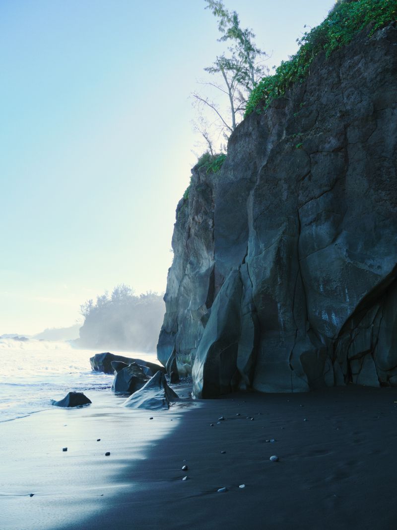 Coastal scene featuring a rugged cliff face covered in vegetation, overlooking a black sand beach and ocean with waves.