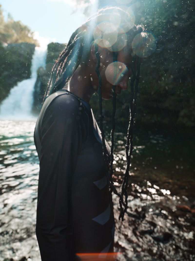 Woman with dreadlocks stands in a river near a waterfall, wearing a long-sleeved shirt. Sunlight filters through the trees.