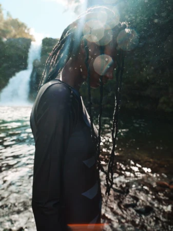 Model Deborah Akanni standing in profile wearing a dark long-sleeve rashguard with braided hair near a scenic waterfall.