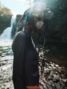Model Deborah Akanni standing in profile wearing a dark long-sleeve rashguard with braided hair near a scenic waterfall.