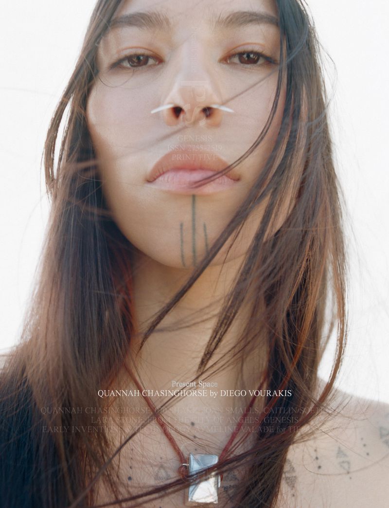 Close-up of an androgynous person with long brown hair, face tattoos and a nose piercing, wearing a pendant necklace against a white background.