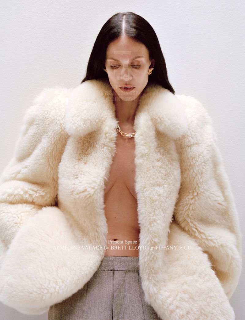 Brunette adult model in a cream-colored oversized fur coat and grey trousers looking down against a white wall in a fashion shot.