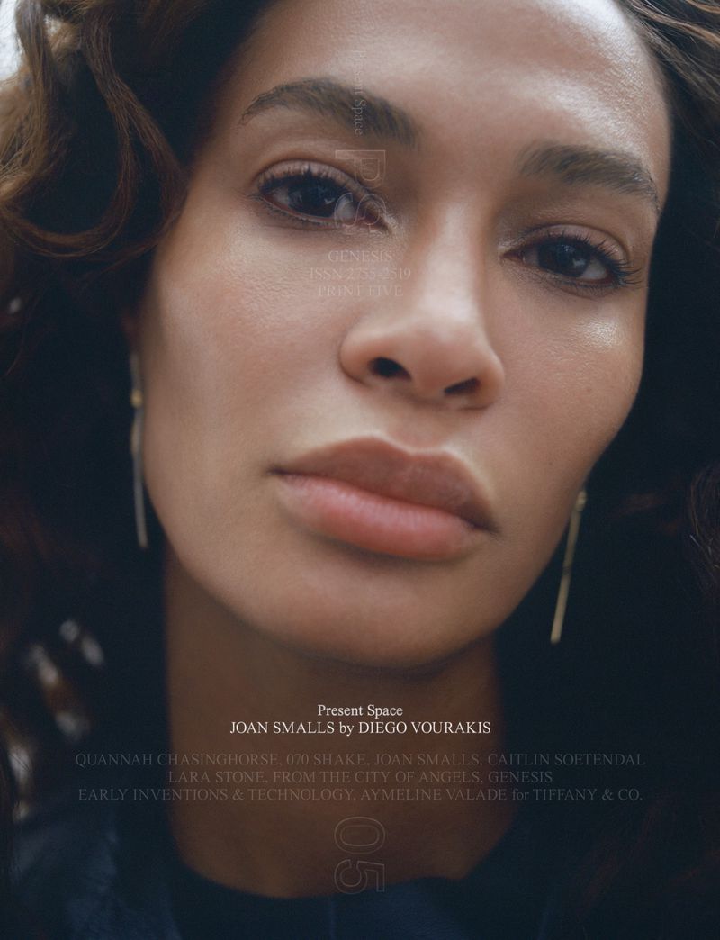 Close-up portrait of Joan Smalls, showcasing her makeup and long, thin gold earrings against a dark background in a beauty editorial shot.