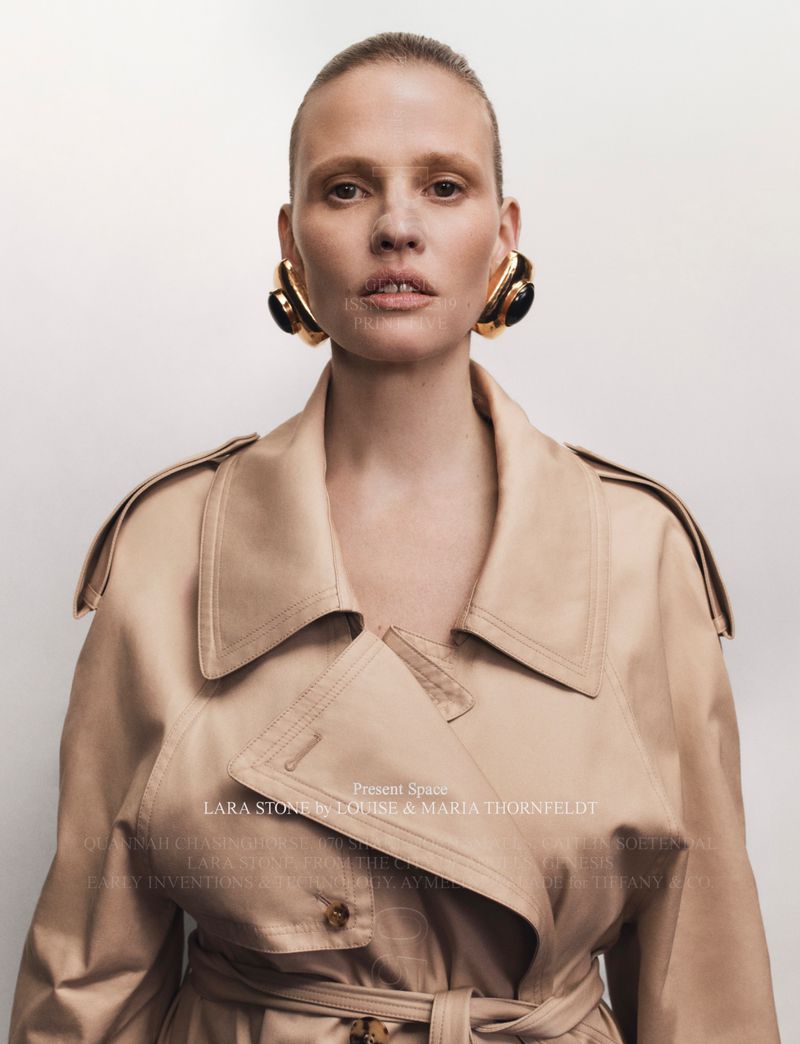 Fashion portrait of an adult female with large gold and black earrings wearing a beige trench coat in front of a white background.