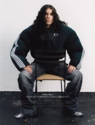 070 Shake seated on a chair wearing a dark puffy jacket and wide denim jeans against a white background