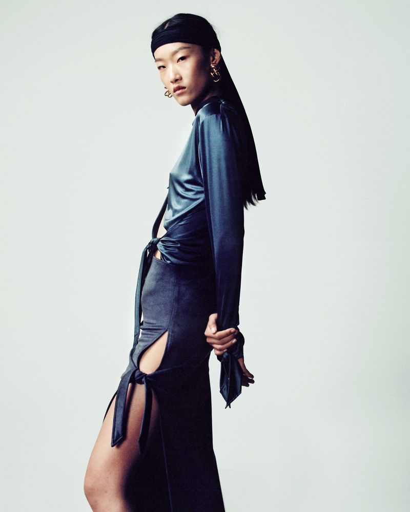 A stylish Asian woman poses in a blue satin wrap top and slit skirt against a clean studio backdrop, showcasing modern elegance.