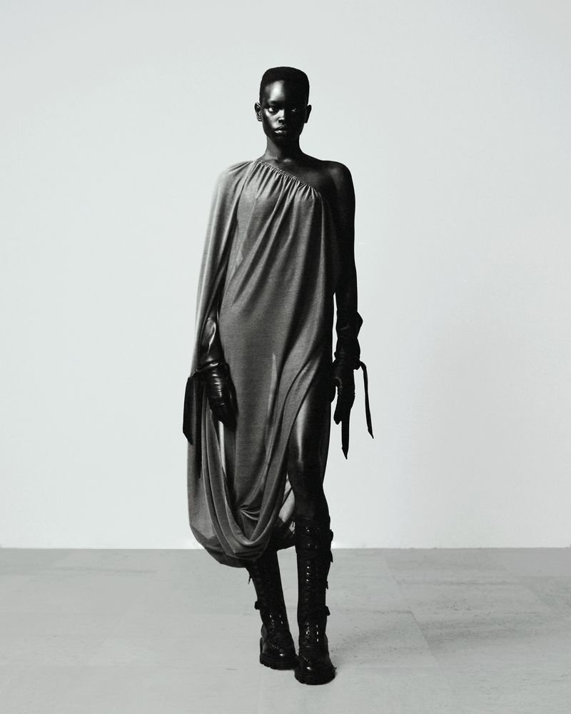 Black and white portrait of a black female model in a draped gray dress, black gloves, and black boots posing against a neutral backdrop.