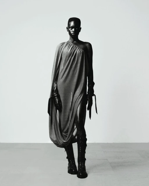 Black and white portrait of a black female model in a draped gray dress, black gloves, and black boots posing against a neutral backdrop.