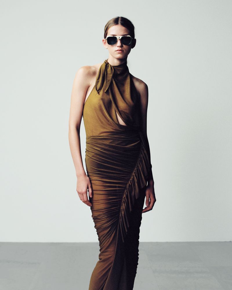 A woman poses in a draped olive-green dress and sunglasses against a neutral backdrop, showcasing a fashion editorial look.