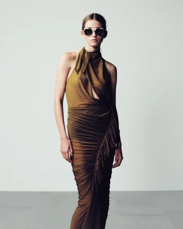 A woman poses in a draped olive-green dress and sunglasses against a neutral backdrop, showcasing a fashion editorial look.