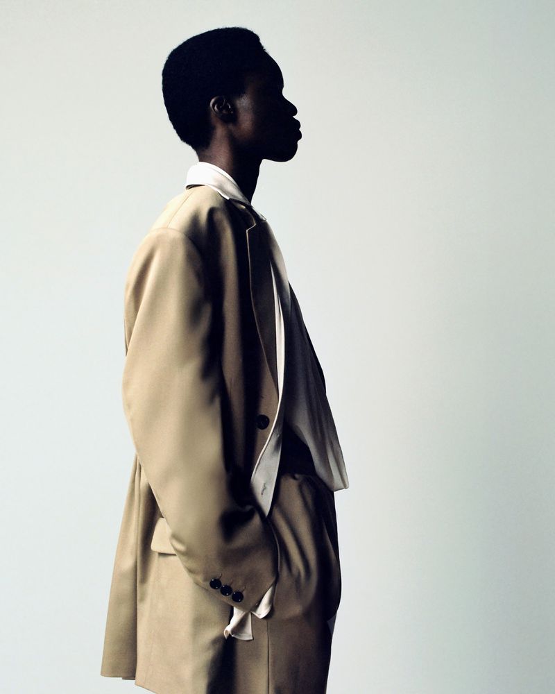Side profile of a model wearing an oversized beige tailored blazer and draped white shirt against a plain light background.