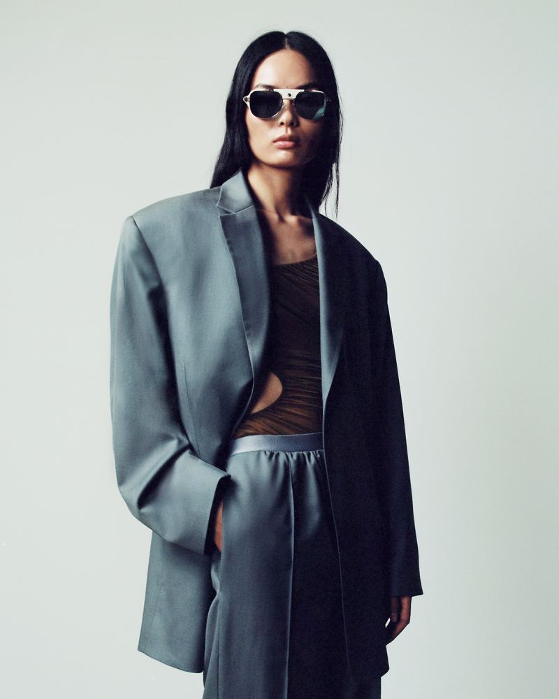 Woman in oversized grey suit and sunglasses poses in a studio setting. Fashion editorial image with cool lighting and minimalist background.