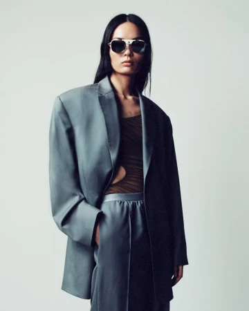 Woman in oversized grey suit and sunglasses poses in a studio setting. Fashion editorial image with cool lighting and minimalist background.