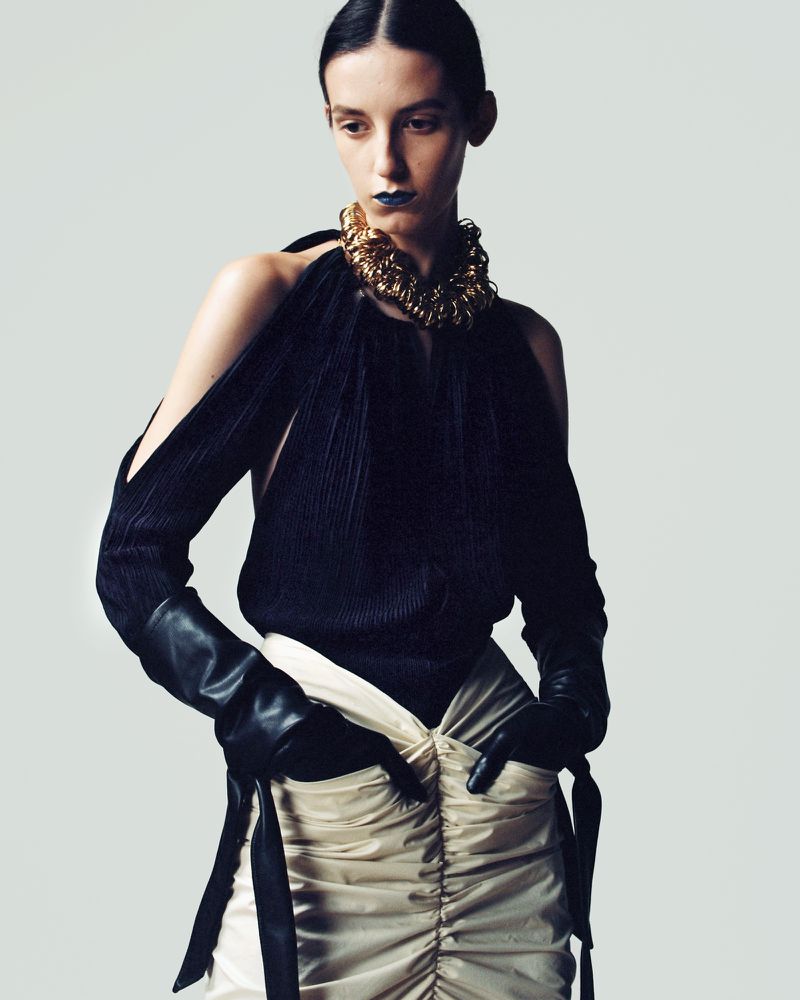 Fashion editorial shot of a woman in a black top, gold necklace, beige skirt, and black gloves posing in a photo studio setting.