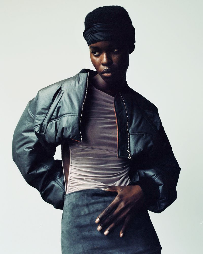 Fashion portrait of an African American woman in a headwrap and leather jacket, posed in a studio with neutral background, looking at the camera.