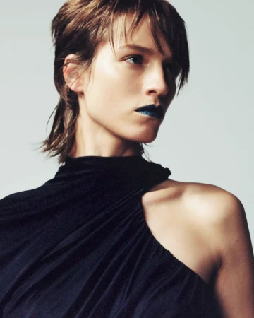 Close-up fashion portrait of a fair-skinned woman with short brown hair and blue lipstick, wearing a black dress against a white backdrop.