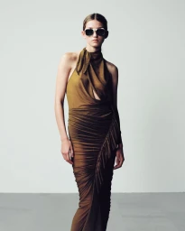 Model wearing a draped brown ruched halter dress and dark sunglasses against a neutral studio backdrop.