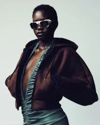 Black model wearing dark futuristic sunglasses, a brown hooded cropped jacket, and a gathered sage green top in a studio setting.