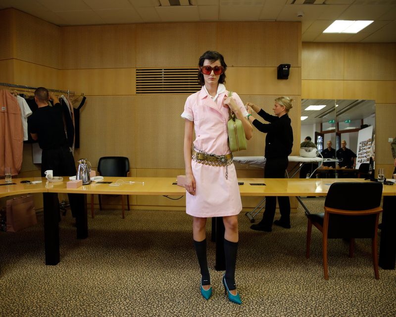 Fashionable woman in a pink dress and red glasses poses in an office, showcasing a vintage style with green bag and retro details.
