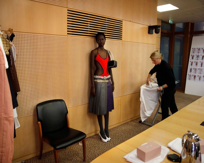 Runway model in cutout swimsuit and skirt posing near a stylist ironing clothing backstage with a neutral indoor background and soft lighting.