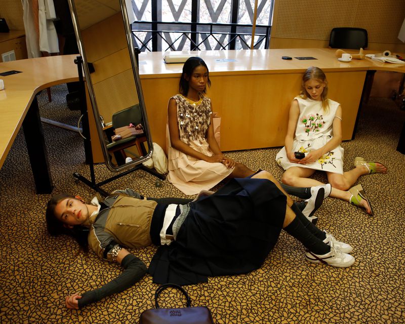 Three fashion models backstage at a runway show, sitting and lying on the floor in dresses, shoes, and sneakers in an indoor setting.