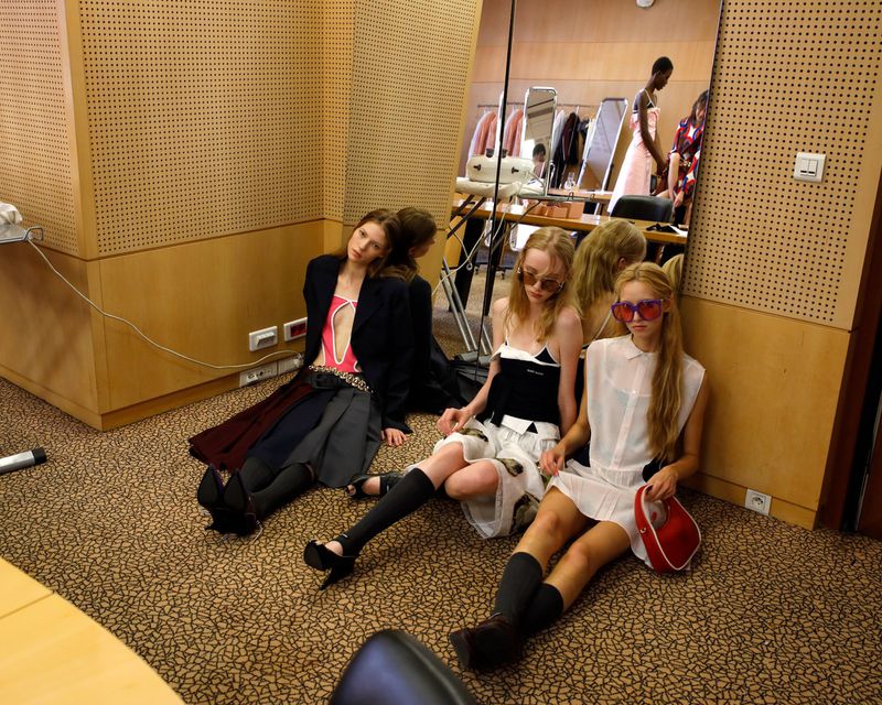 Three fashion models are sitting on the floor in a backstage setting. The models wear trendy outfits and pose in front of a mirror.