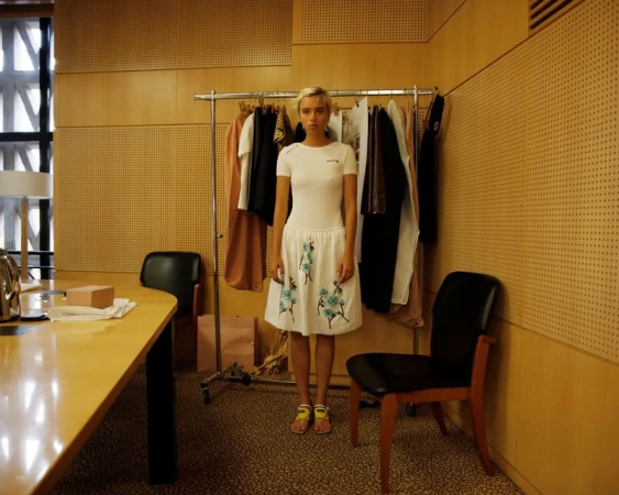Model in a white short-sleeve dress with blue floral embroidery standing in front of a clothing rack in a wood-paneled room.