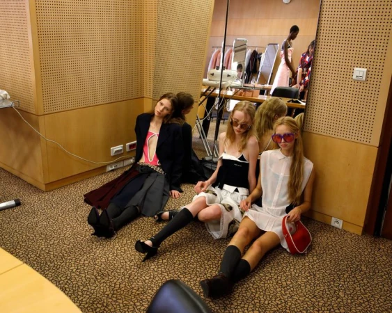 Three models wearing designer dresses, sunglasses, and knee-high socks sit on a carpeted floor in a backstage dressing room.