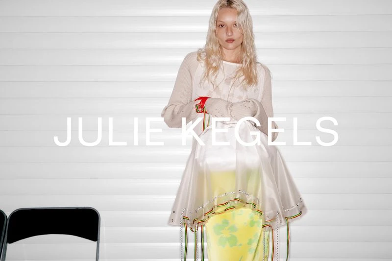 Young woman wearing a light beige sweater and a yellow and white skirt stands in front of a textured white wall.
