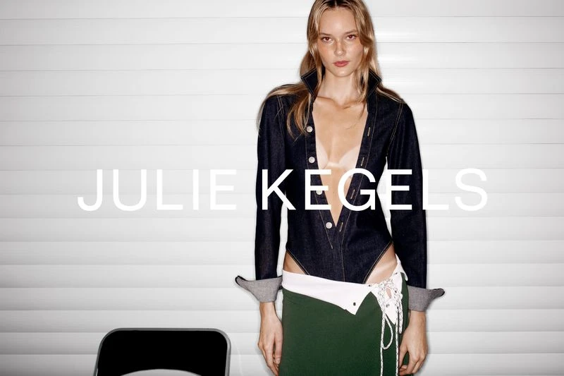 Fashion shot of a woman posing in a denim jacket and green skirt in front of a white wall with the name 'JULIE KEGELS' on the wall.