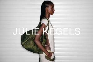 Black female model in profile wearing a cream tunic and holding an oversized olive green bag in front of a white slatted wall.