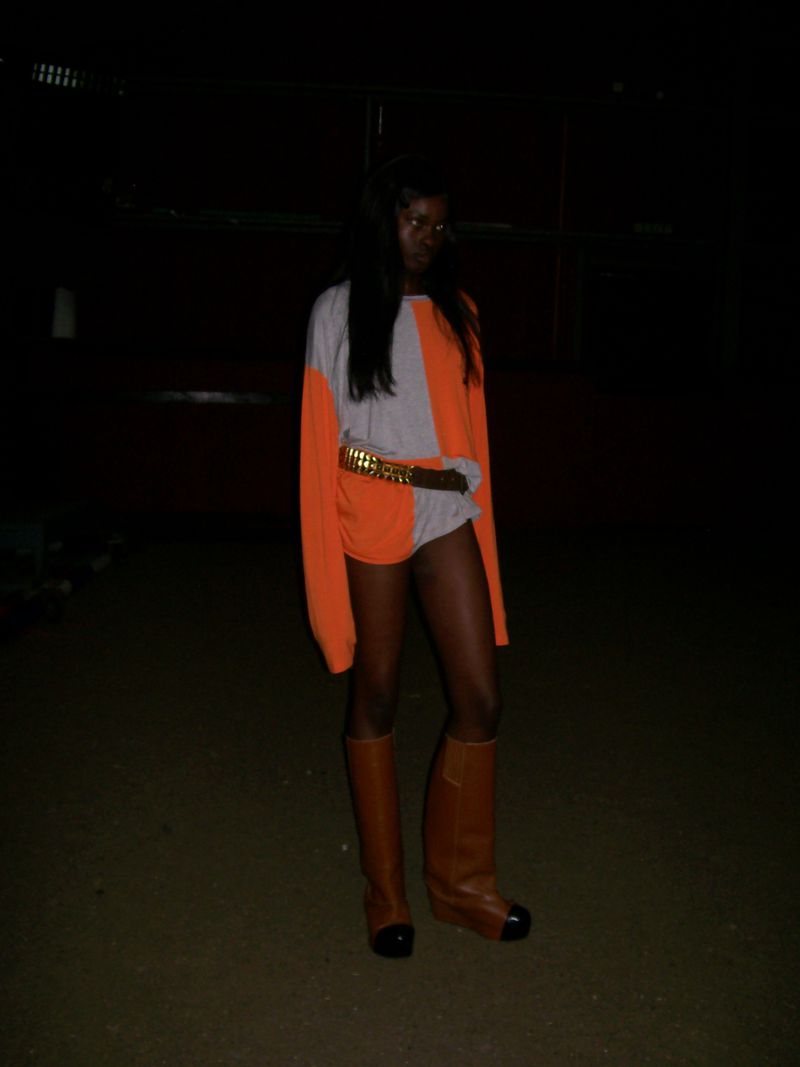 A full-length shot of a woman in a color-block sweater dress with a gold belt and high boots, posing in a dimly lit outdoor setting at night.