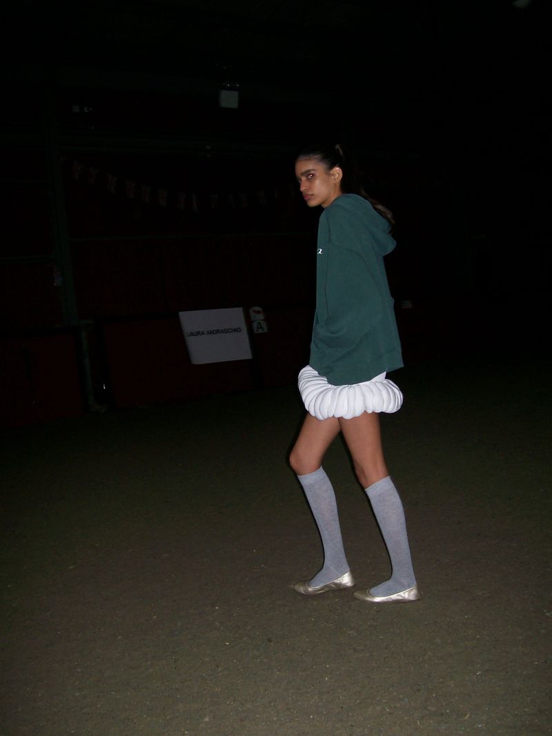 Young woman wearing a green hoodie, white skirt, and gray socks stands outside in a dark environment looking at the camera.